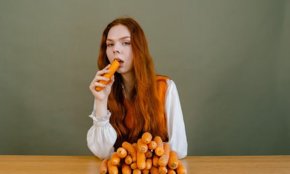 Red-haired woman sits at a table, holding a carrot, surrounded by a stack of vegetables.